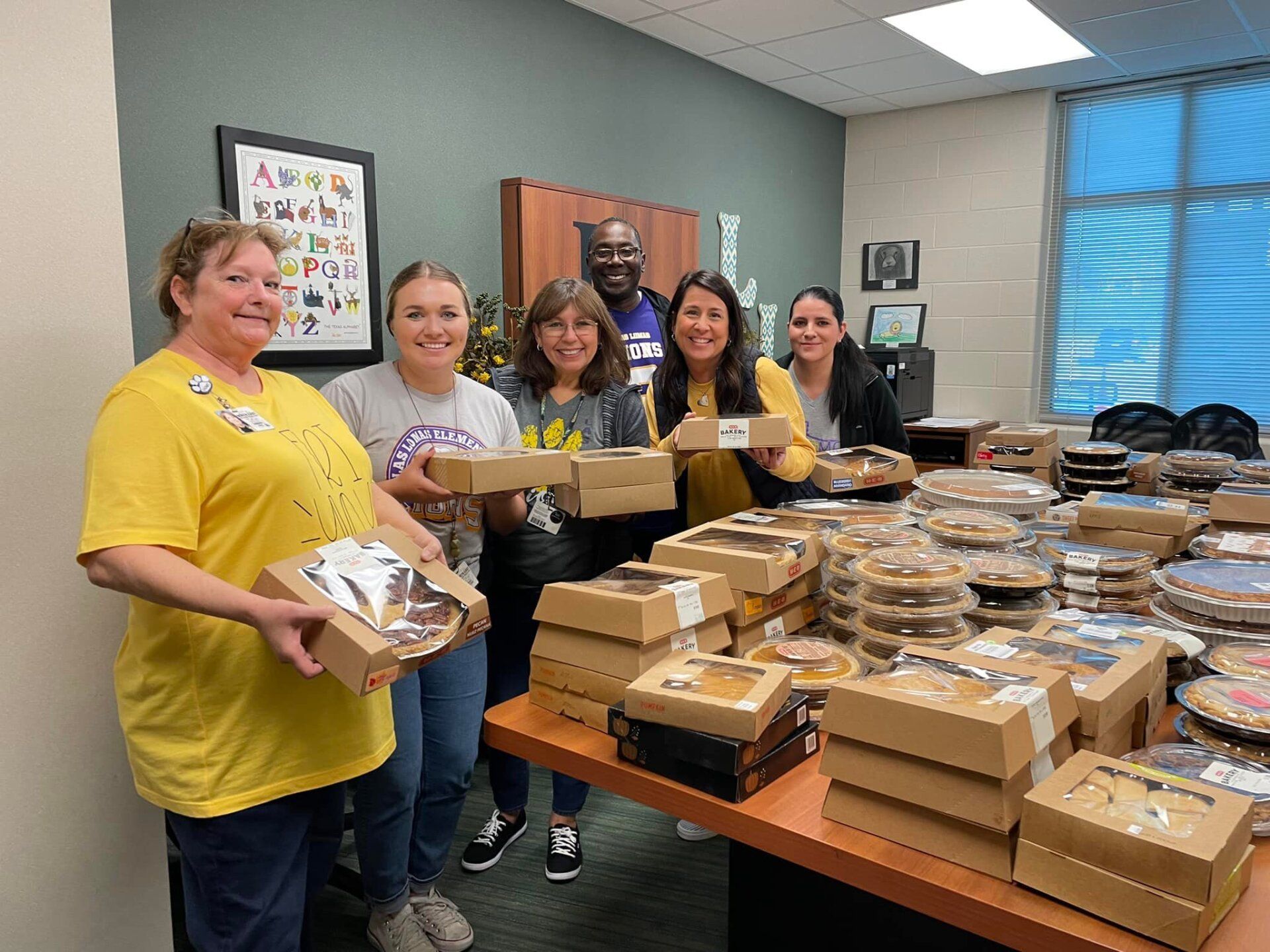 Five women in an office smile beside tables stacked with boxed meals and trays, preparing a food distribution.