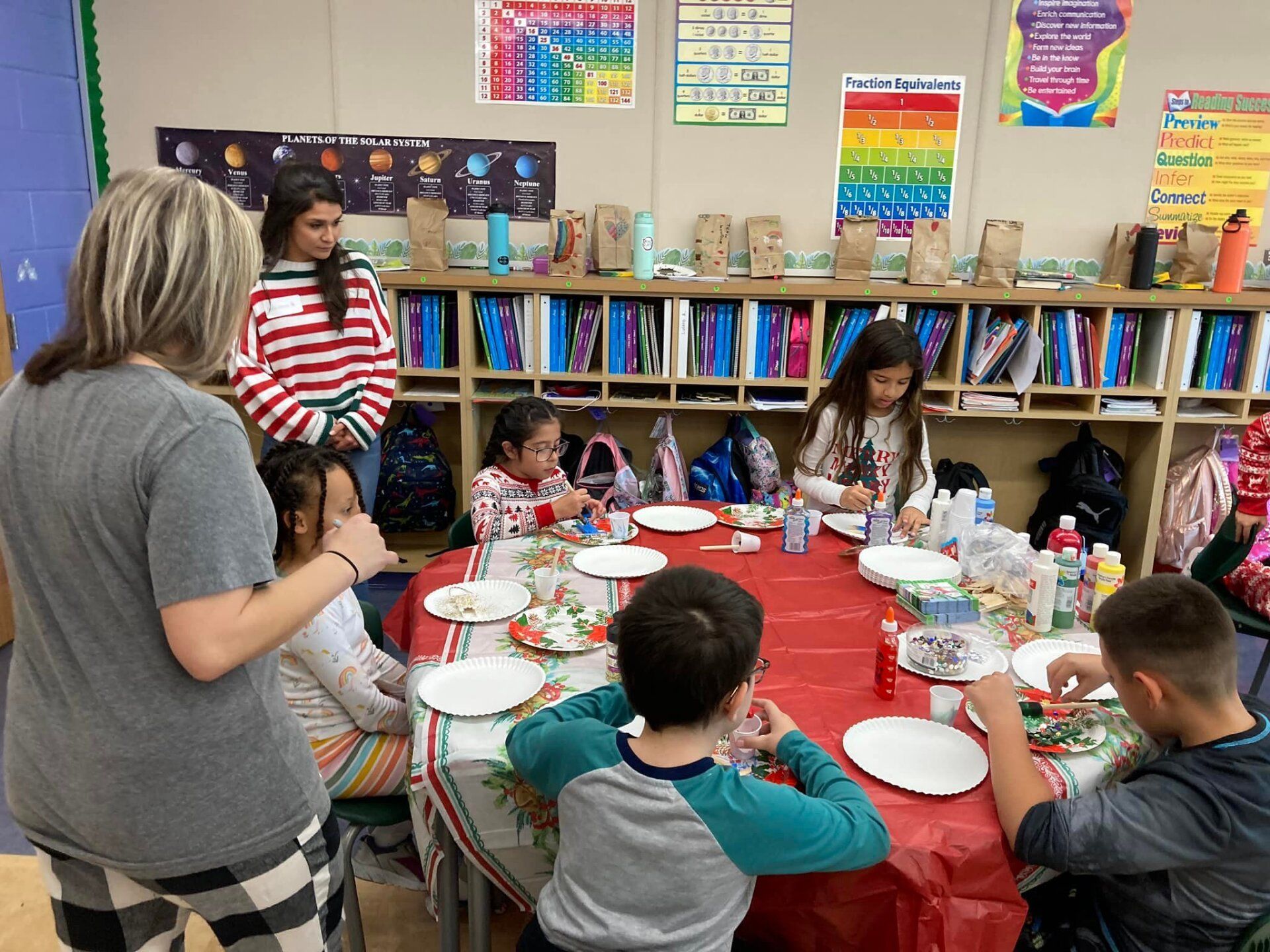 Children seated at a red-covered table in a classroom, making crafts with an adult nearby.