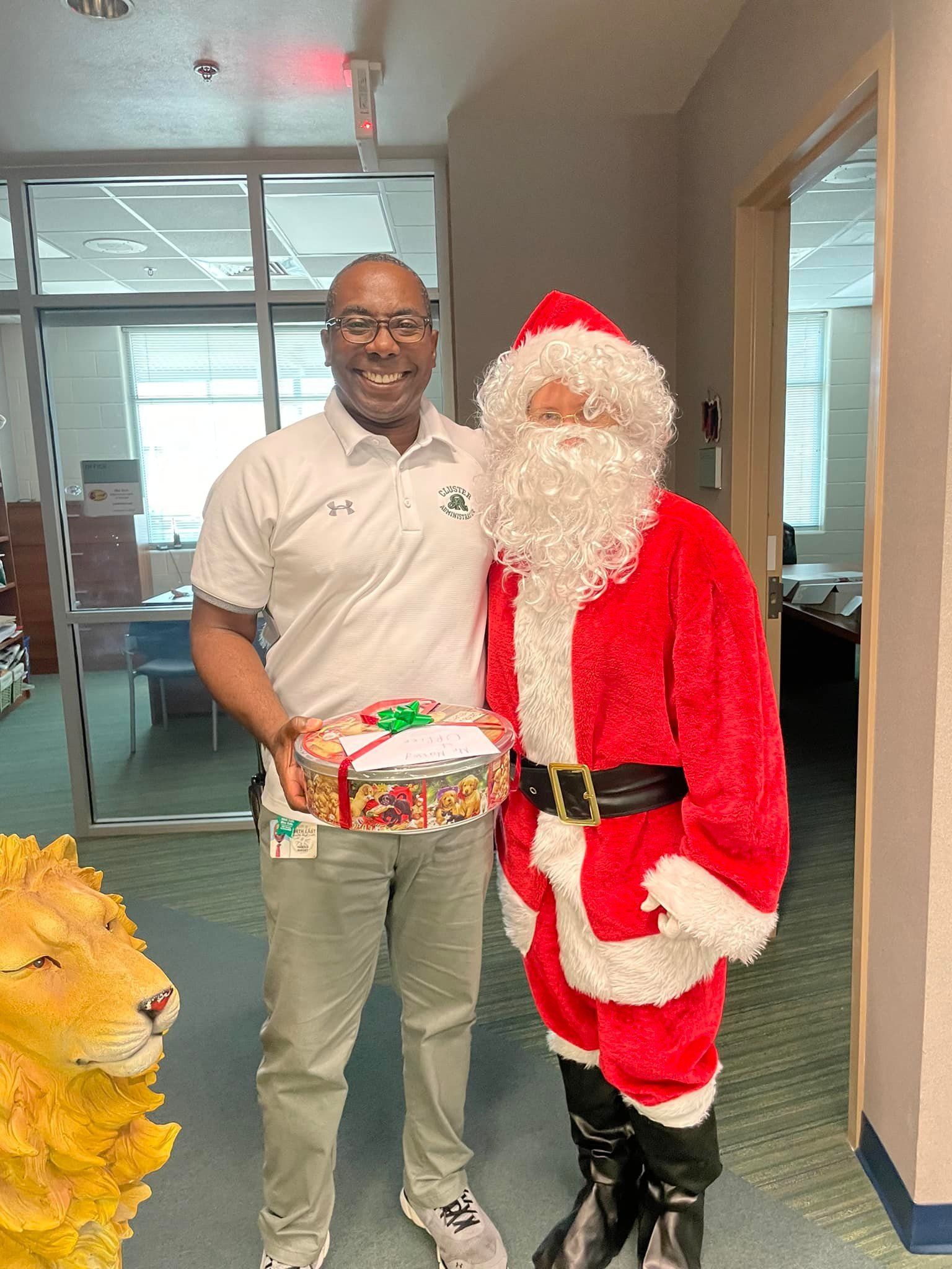 Man standing beside Santa in an office hallway, holding a wrapped gift.