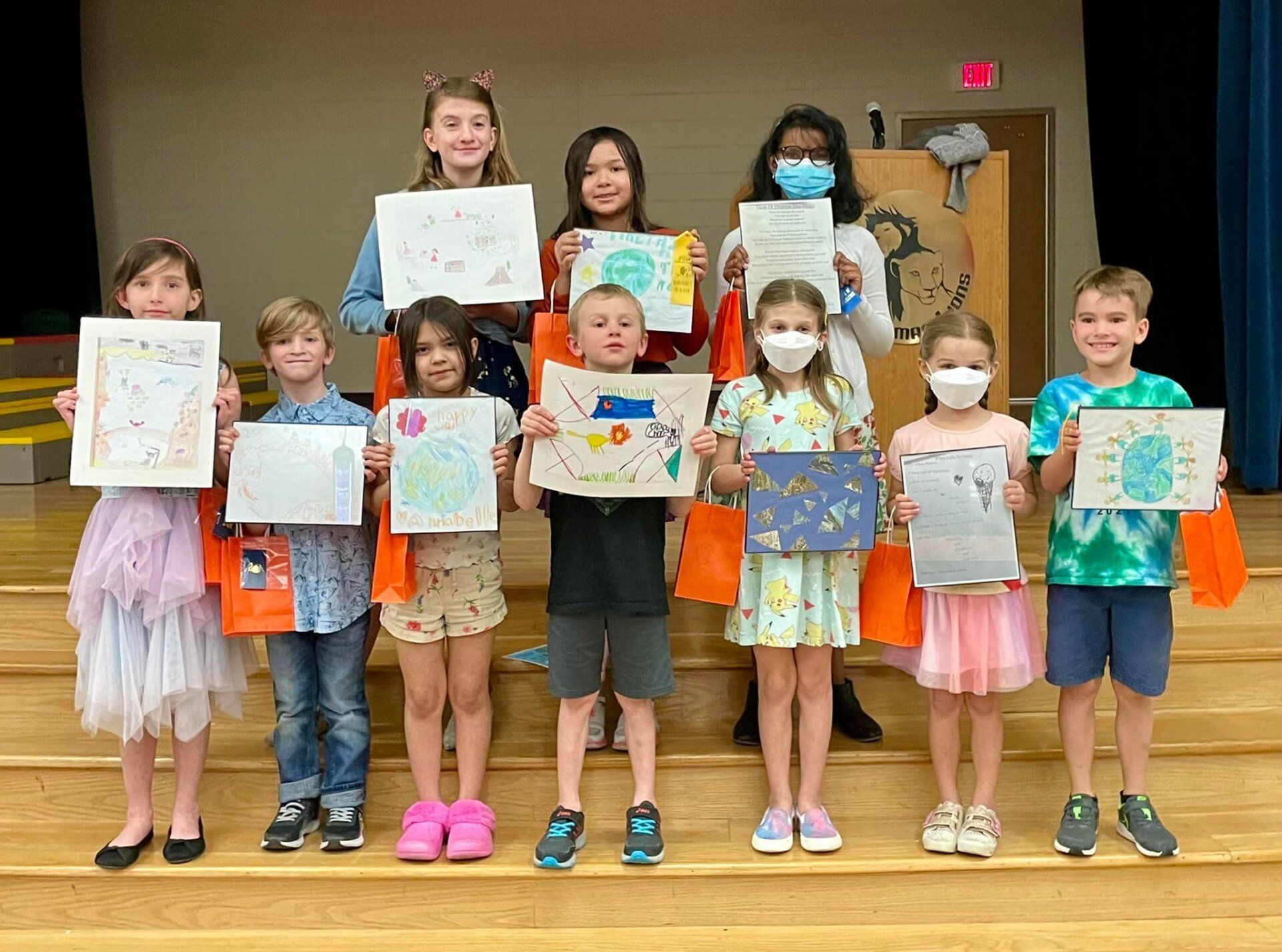 Children standing on a stage holding colorful artwork and certificates, smiling for a group presentation