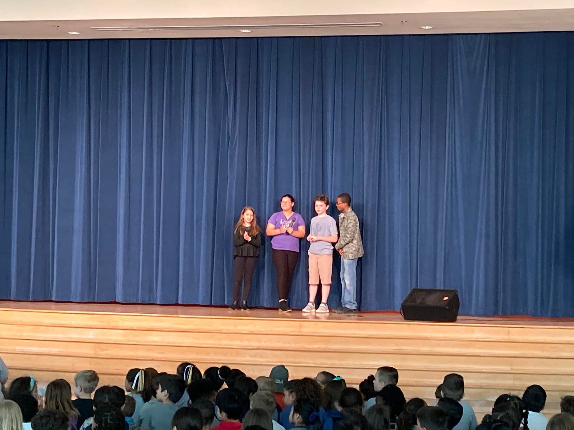 Four people stand on a stage before blue curtains, facing an audience.