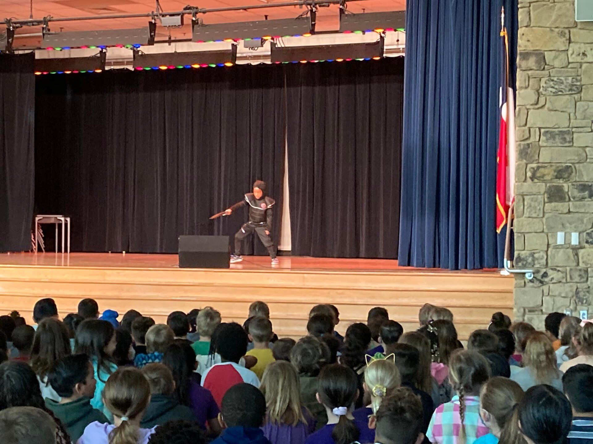 Person speaking on a school stage before an audience, with black curtains and a blue curtain at the side.