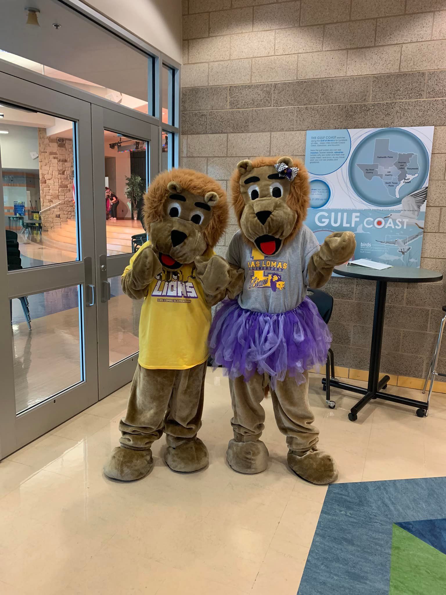 Two mascot characters in yellow shirts stand indoors beside a Gilead sign.