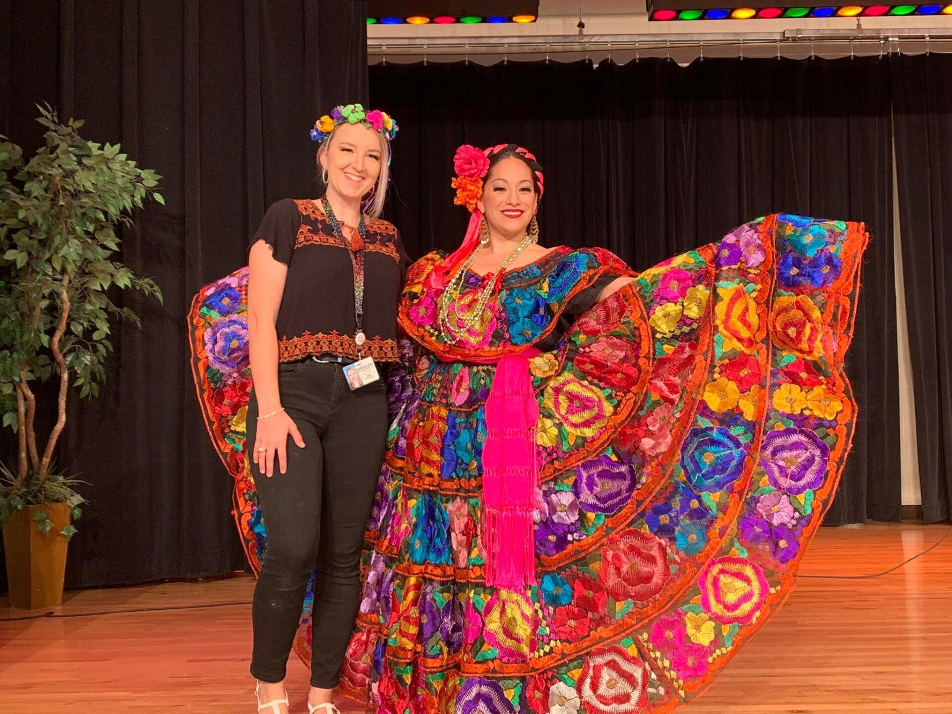 Two women posing on a stage, one in black and one in a colorful traditional dress with a wide floral skirt