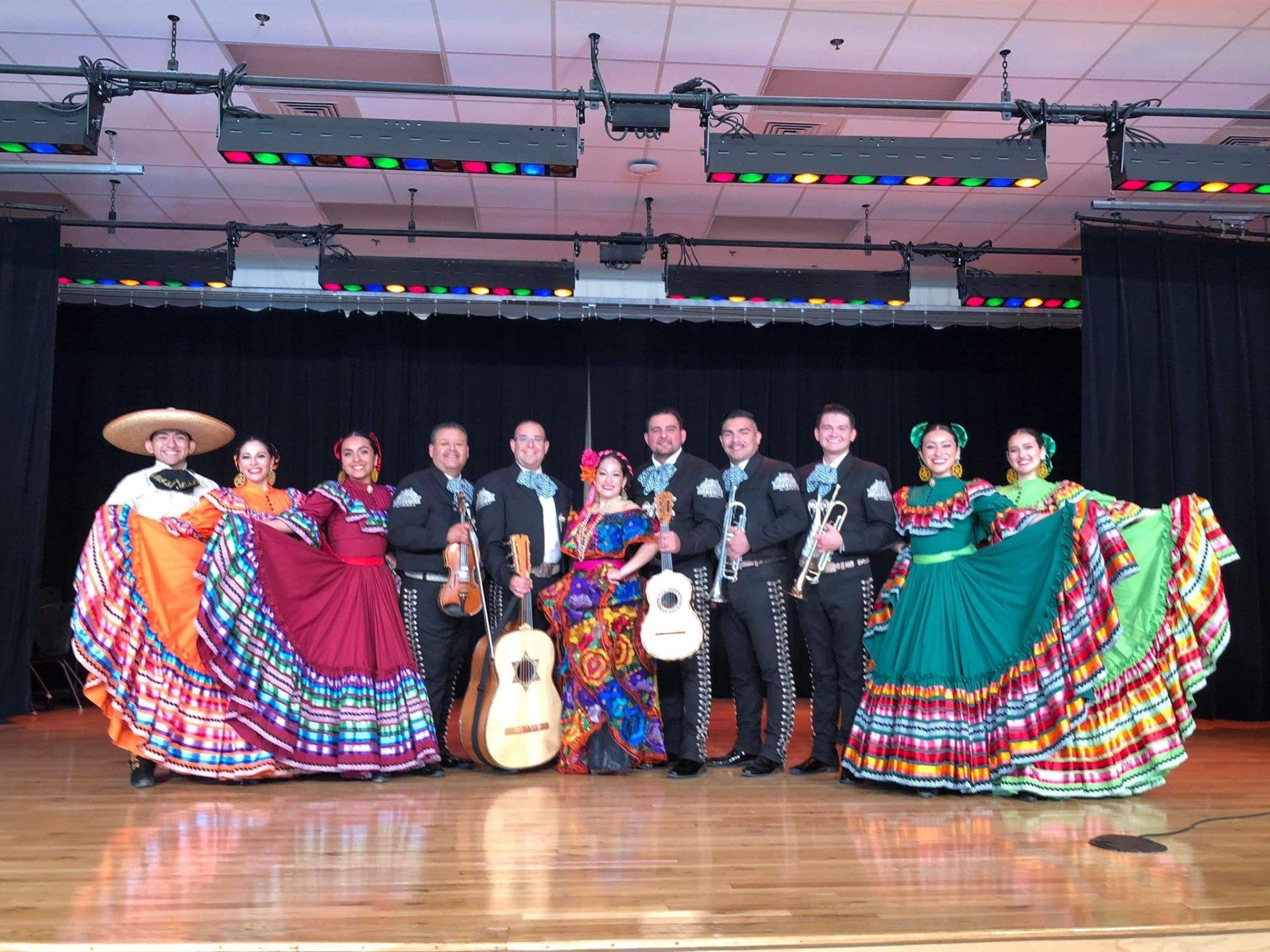 Mariachi band in black suits onstage with colorful dancers in traditional dresses and sombrero