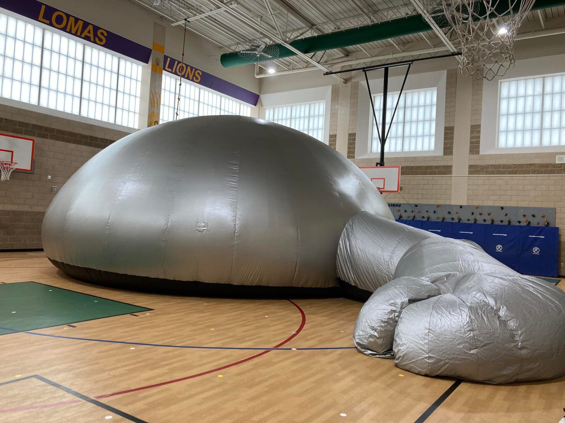 Large silver inflatable dome in a gymnasium, with a smaller attached inflatable section on the floor.
