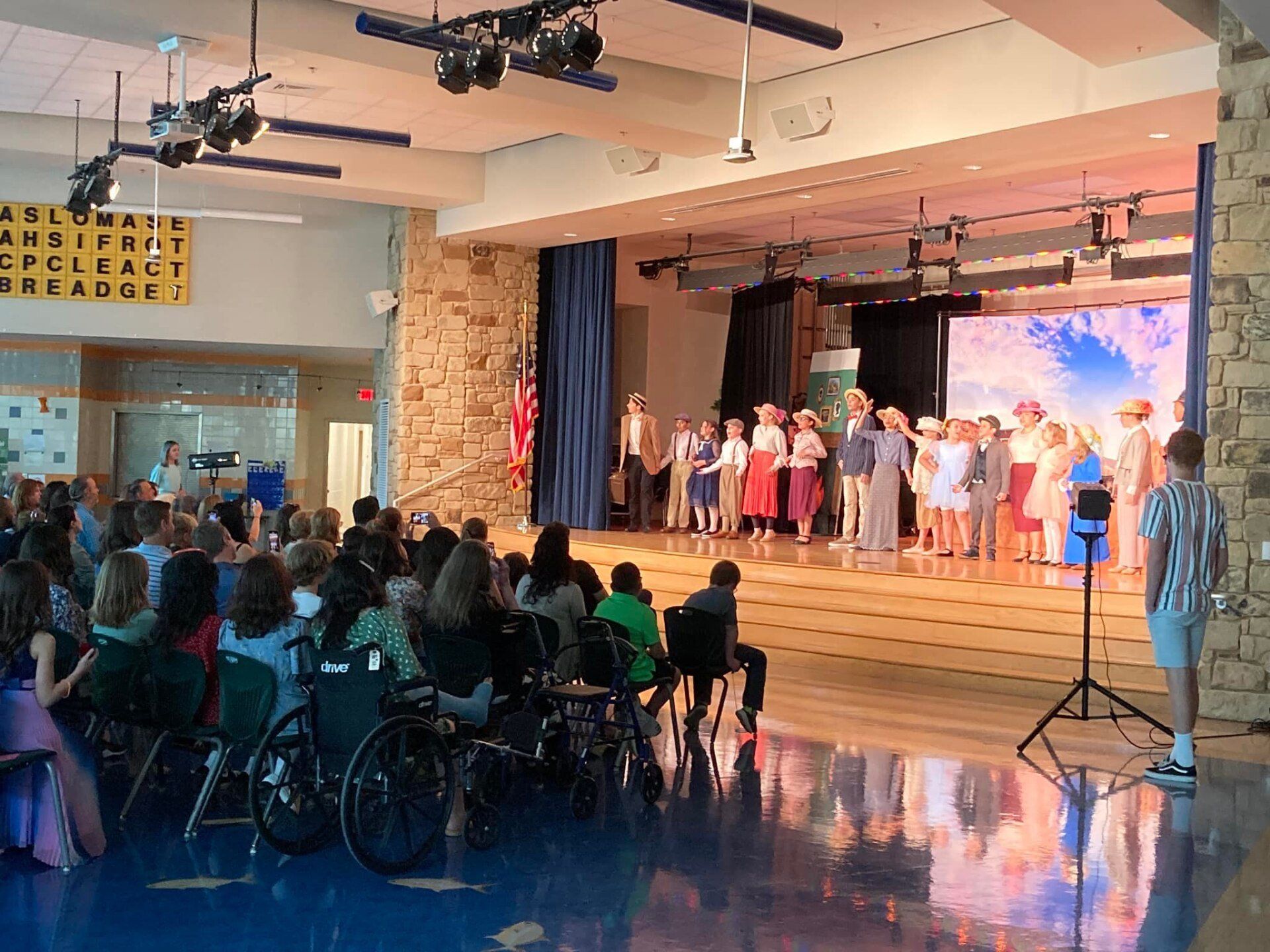 Children performing on a lit stage in a school auditorium, with audience seated in front