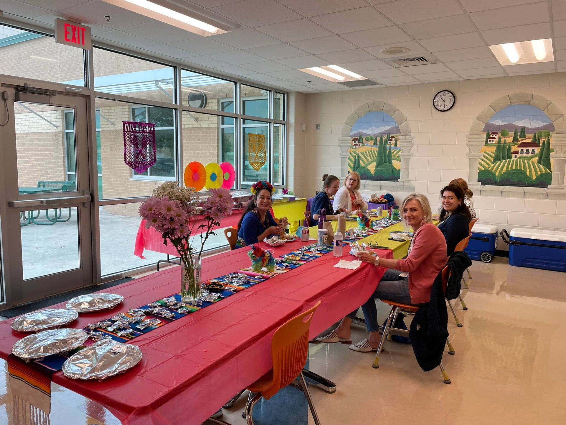 People seated at a decorated banquet table in a bright community room with murals and balloons.