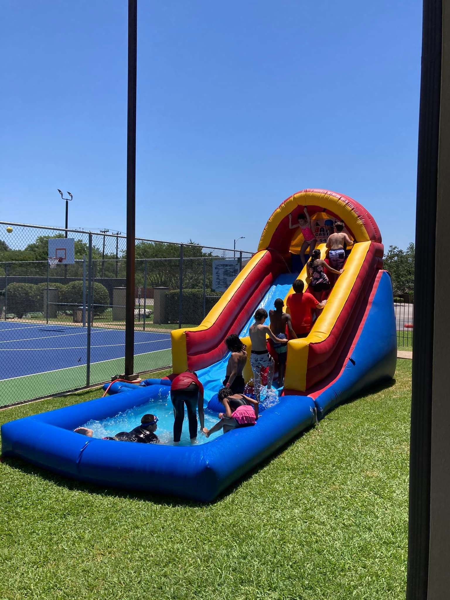 Children playing on a colorful inflatable water slide and splash pool on a sunny grassy field.