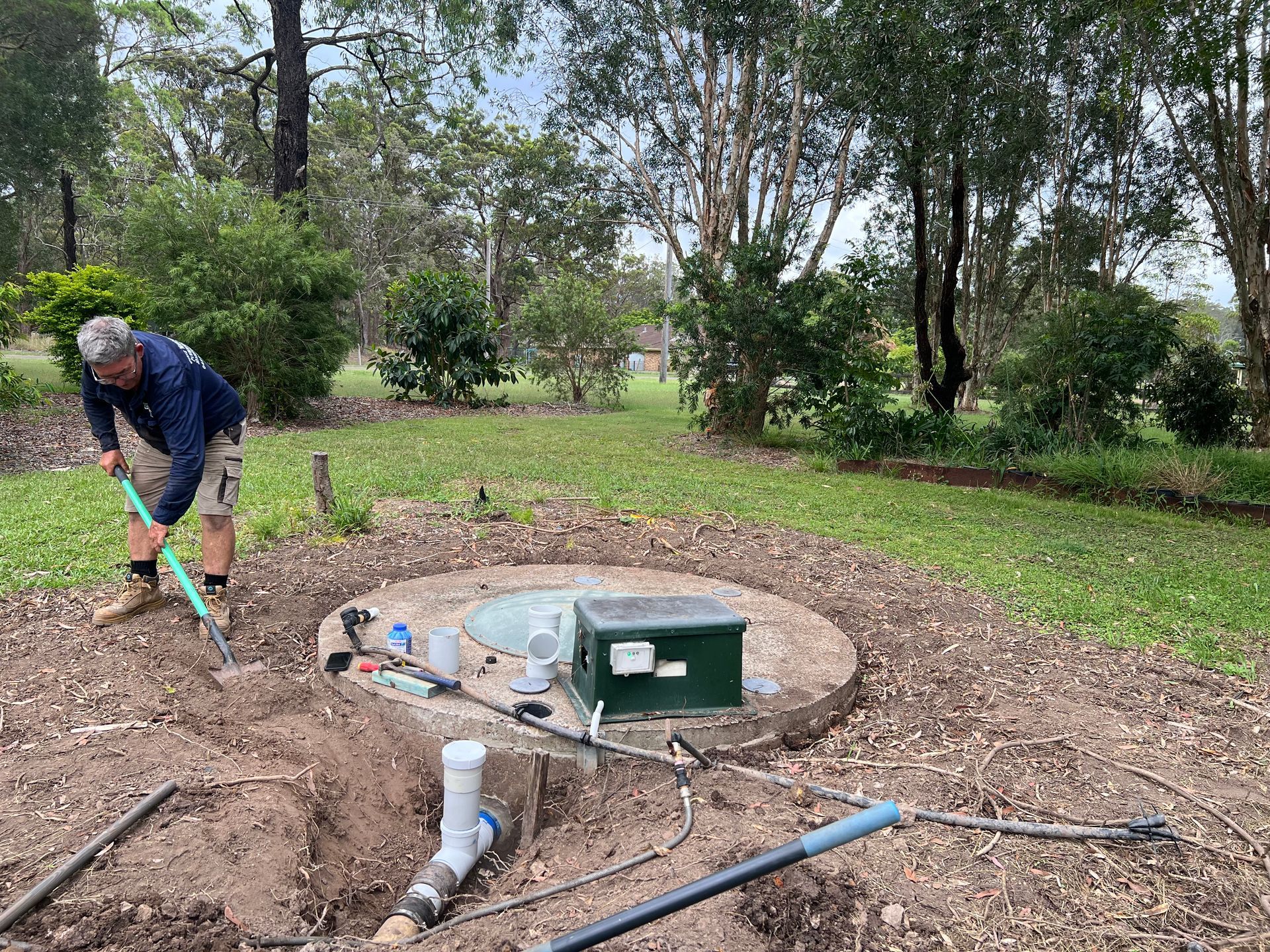 A Person in A Blue Shirt Uses a Shovel to Clear Dirt Near an Outdoor Utility — Chris Murphy Plumbing and Maintenance in Langley Vale, NSW