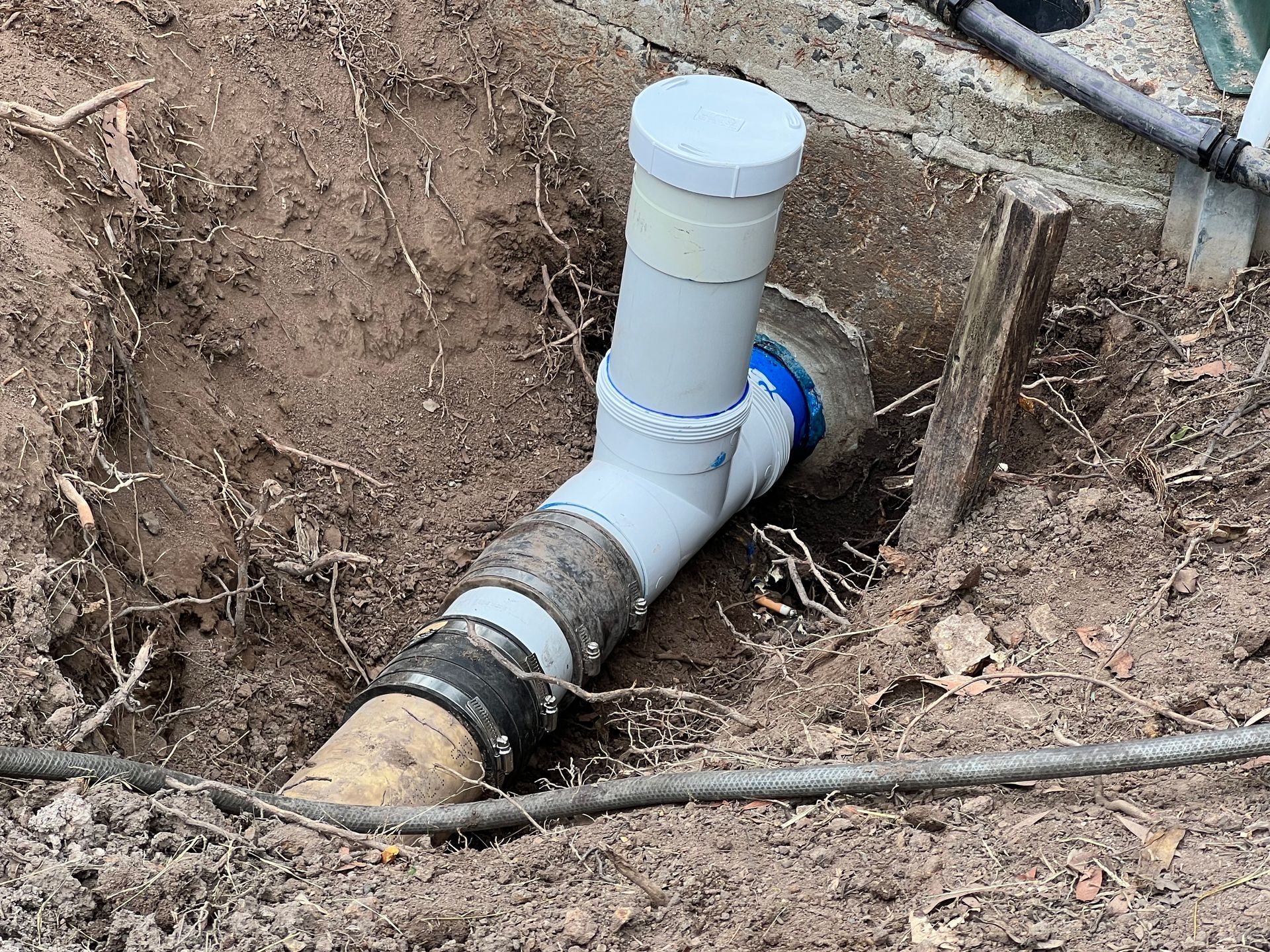 A white PVC T-pipe cleanout installed in a trench, connecting to an existing pipe with black rubber couplings.