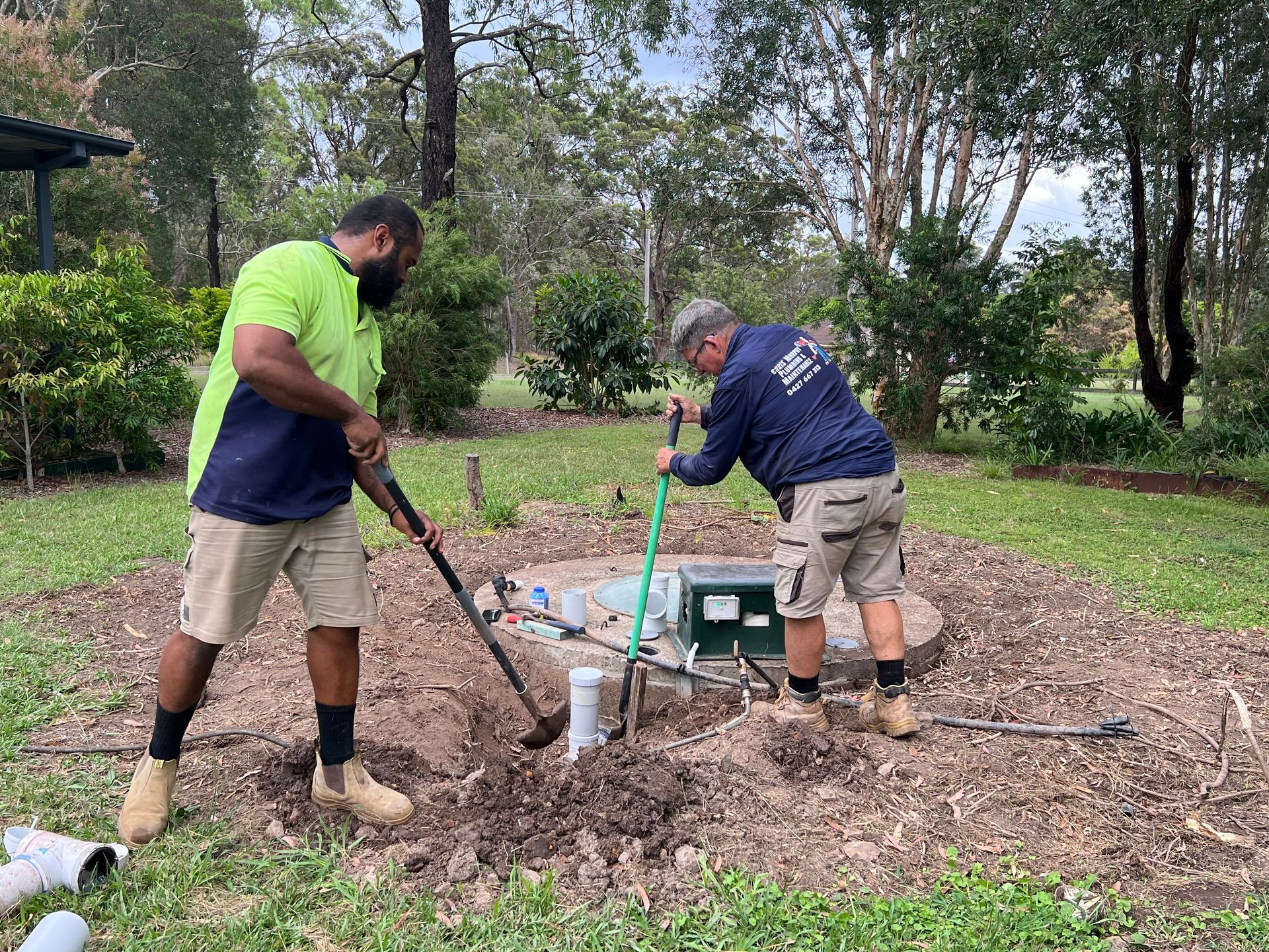 Two people use shovels to work on an outdoor landscape project near a green utility box and a small white pipe.