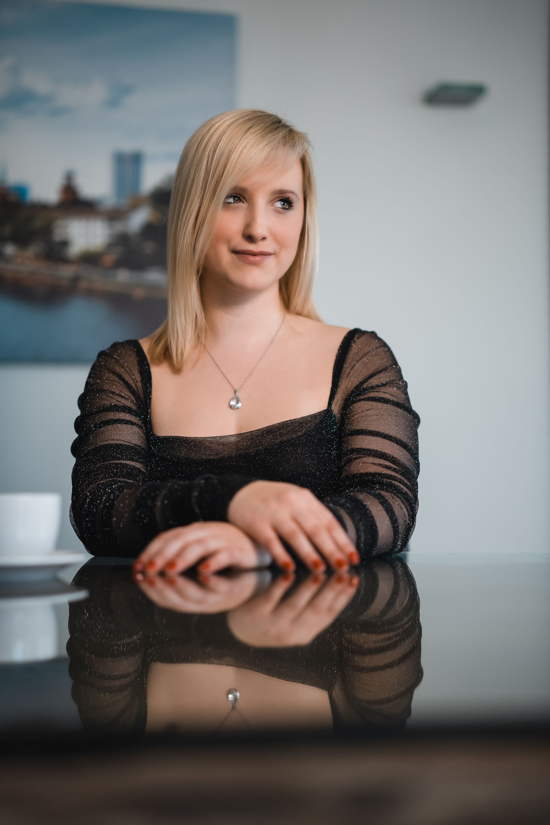 Blonde Frau in schwarzem Perlentop sitzt an einem Hochglanztisch und blickt ins Off. Im Hintergrund eine weiße Tasse.