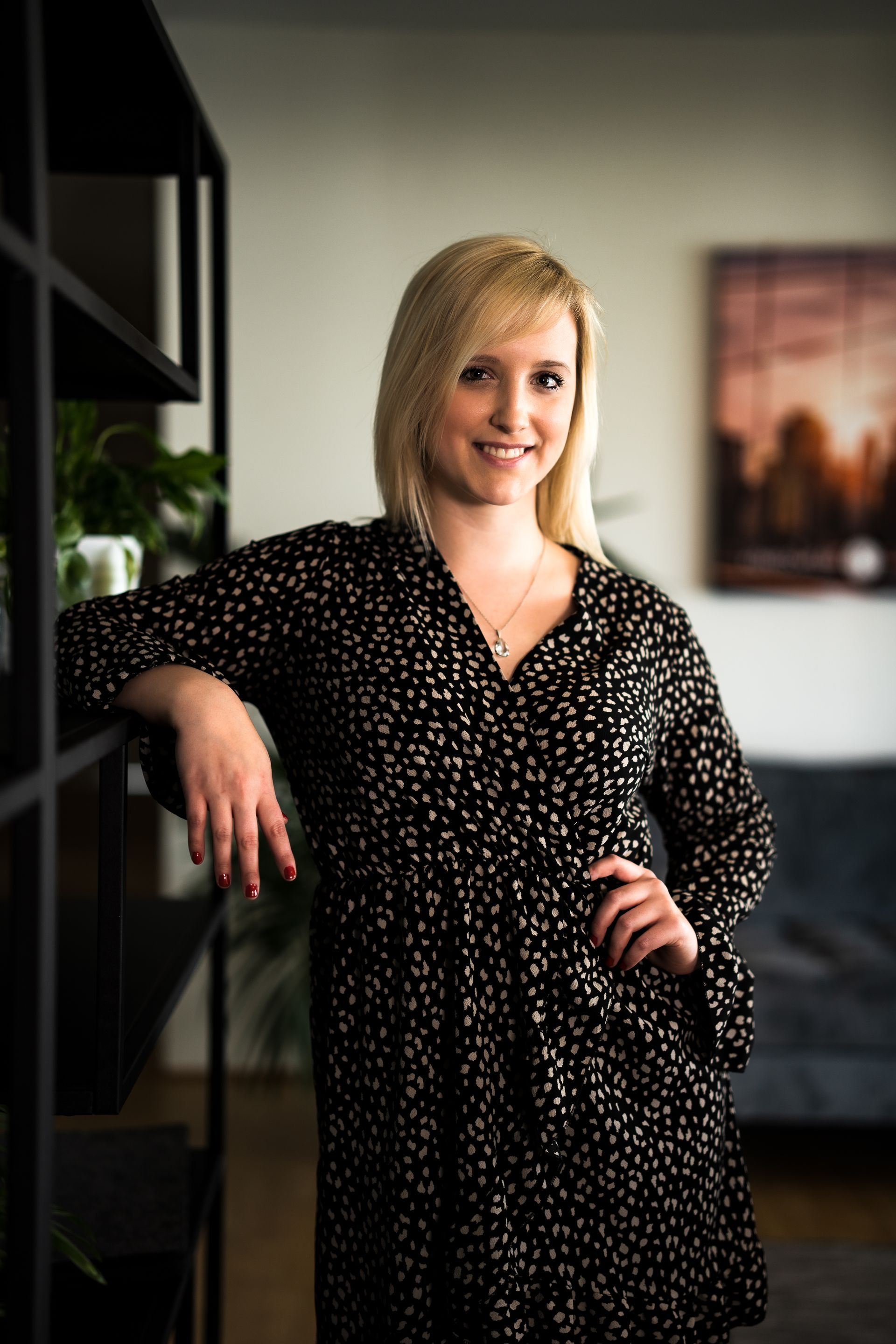Blonde Frau im schwarz gepunkteten Kleid lächelt und lehnt drinnen an einem schwarzen Bücherregal.