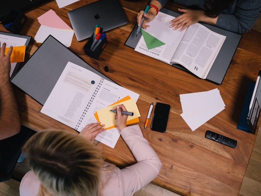 Menschen lernen mit Stiften und Markern an einem Holztisch mit Büchern, Laptops und Notizen.