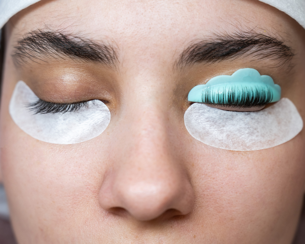 A person's face with a lash lift in progress. One eye is closed with a protective pad and lashes, while the other has a lash lift tool applied.
