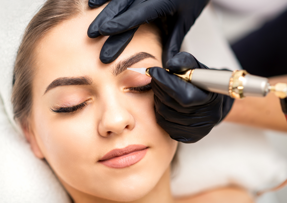 Woman receiving an eyebrow tattoo with eyes closed.  A gloved hand holds a tattooing machine, performing the procedure in a salon.