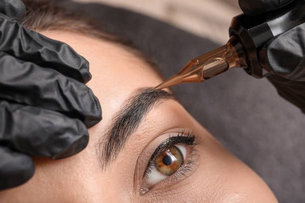 Eyebrow tattooing in progress; gloved hands, machine, and brown ink near eye.