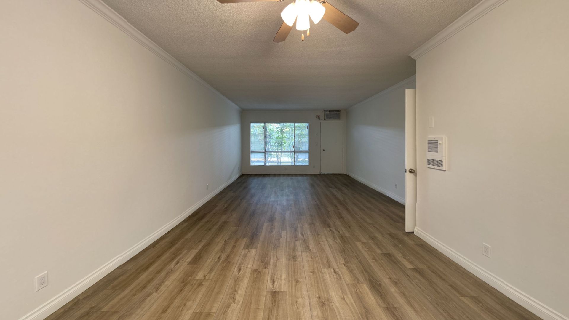 Empty room with wood-look floor, white walls, large window, ceiling fan, and open door.