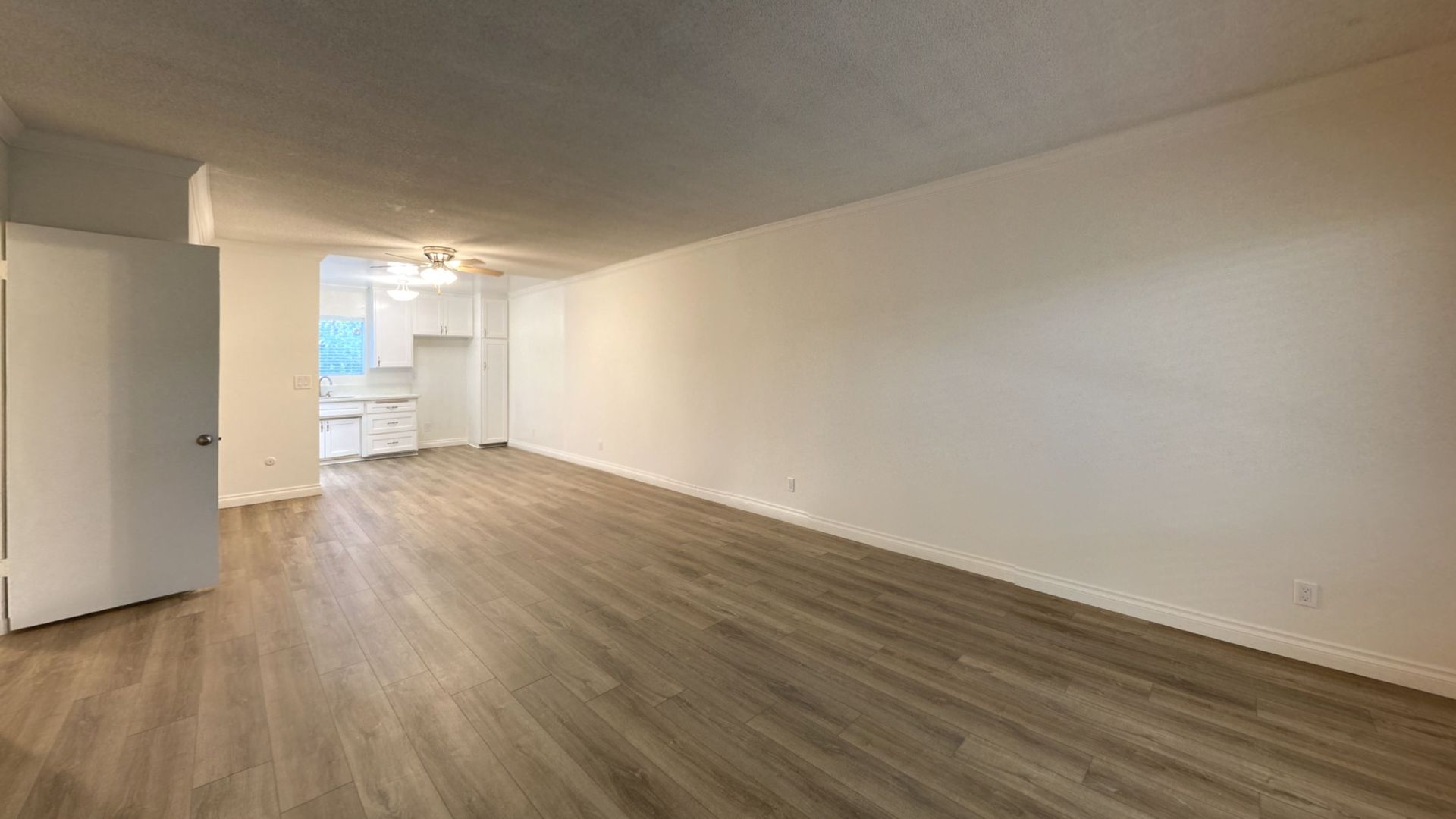 Empty living room with wood-look flooring, white walls, and a doorway leading to a kitchen.