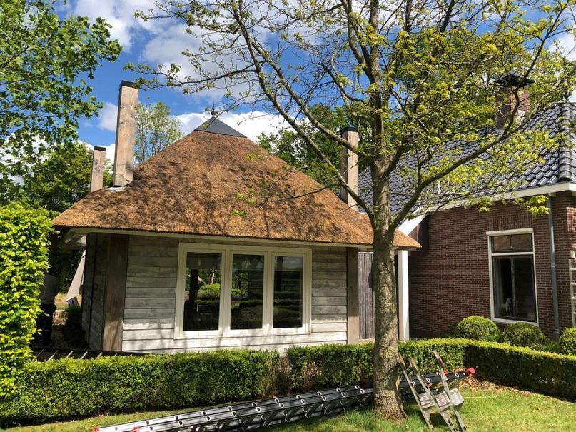 Klein huisje met een rieten dak en grote ramen, naast een bakstenen gebouw.