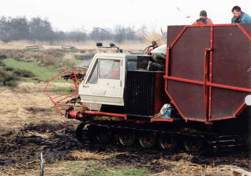 Een rupsvoertuig met een grote rode container in een moerassig gebied. Twee mensen zitten in de container.