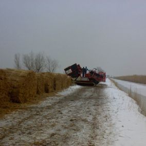 Rode machines op een besneeuwde zandweg naast rietbalen. Twee mensen staan bij de gekantelde machine op een bewolkte dag.