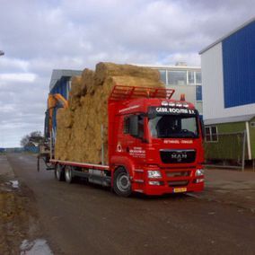 Rode vrachtwagen vervoert rietbalen over de weg, naast gebouwen, onder een bewolkte lucht.