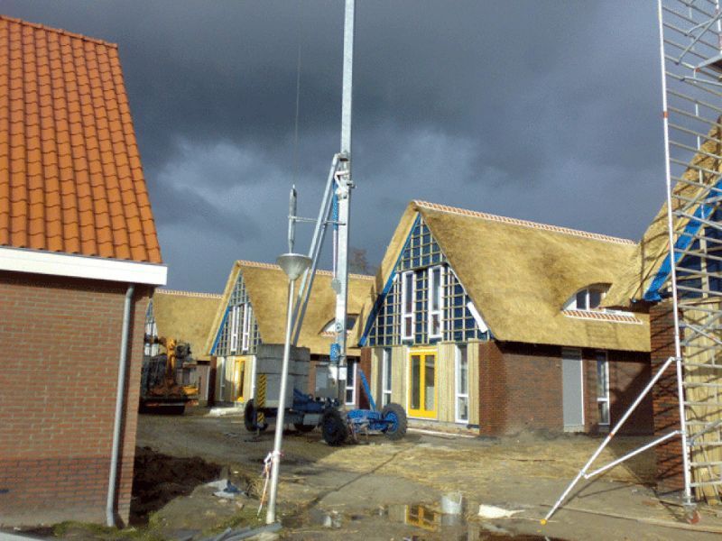Huizen in aanbouw met rieten daken en steigers; blauwe lift op de voorgrond. Bewolkte lucht.