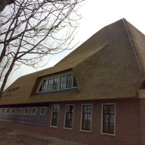 Een bakstenen gebouw met een groot rieten dak. Er zijn ramen zichtbaar. Op de achtergrond staat een kale boom.