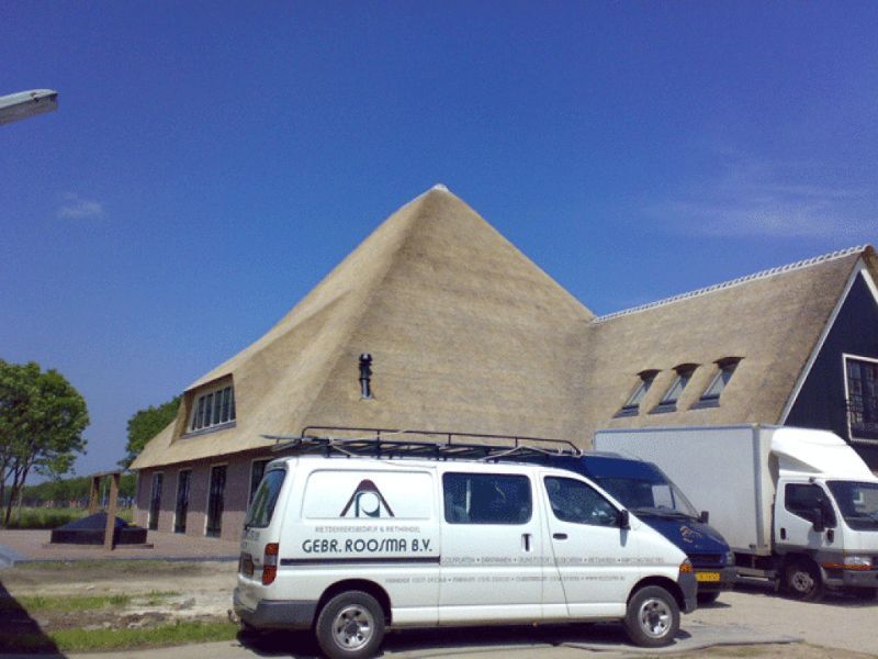 Witte bestelwagen met bedrijfslogo geparkeerd bij een gebouw met een rieten dak. Persoon aan het werk op het dak.