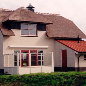 Wit huis met een rieten dak, rode ramen en een kleine rode deur, afstekend tegen een bewolkte lucht.