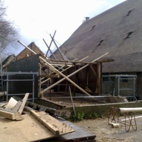 Bouwplaats met houten framewerk voor een gebouw met rieten dak; bouwmaterialen liggen verspreid.
