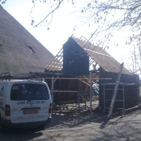 Een rieten dak in aanbouw, naast een afgebouwd dak. Een witte bestelwagen staat vlakbij geparkeerd.