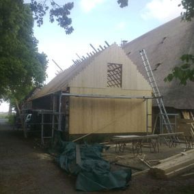 Schuur in aanbouw; steigerwerk, houten gevelbekleding, rieten dak zichtbaar.