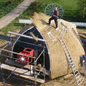Man bedekt het dak van een windmolen met riet, met behulp van steigers en een ladder.