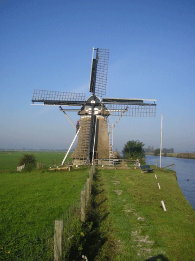 Windmolen op een groene, grazige oever, naast een kanaal. Blauwe lucht op de achtergrond.
