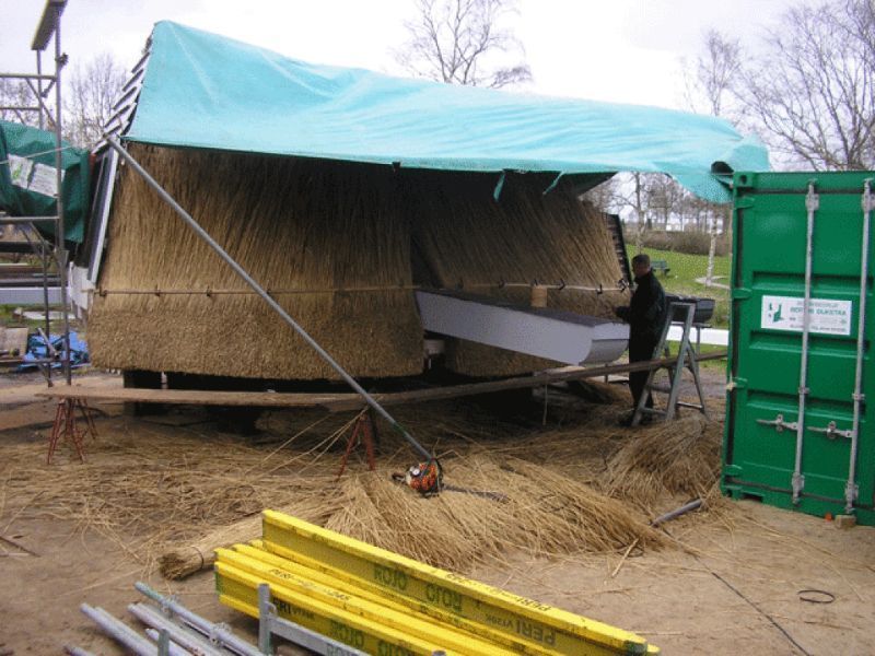 Constructie van een rieten dak: persoon aan het werk, ondersteund door steigers, groen zeil erboven, container in de buurt.