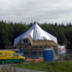 Rond gebouw in aanbouw, gedeeltelijk bedekt met zilverkleurig materiaal, met details van een rieten dak en bouwmaterieel.