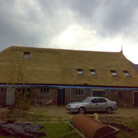 Schuur met rieten dak, kleine ramen en een zilveren auto ervoor geparkeerd. Bewolkte lucht.
