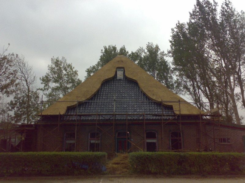 Schuur met rieten dak, gevel met donkere tegels en steigerwerk. Bewolkte dag.