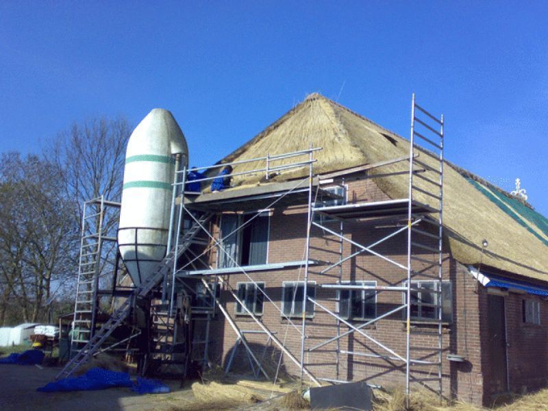 Gebouw in aanbouw met een rieten dak, steigers en een grote siloconstructie tegen een helderblauwe lucht.