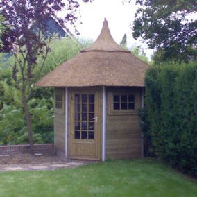 Houten tuinhuisje met rieten dak, gelegen in een groene tuin met haag en bomen.