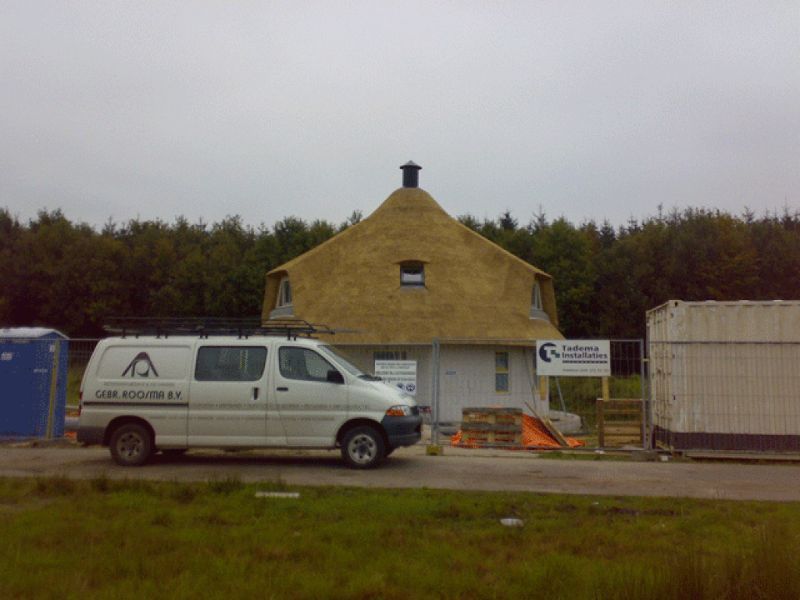 Witte bestelwagen geparkeerd voor een rond huis met een rieten dak, bouwplaats.
