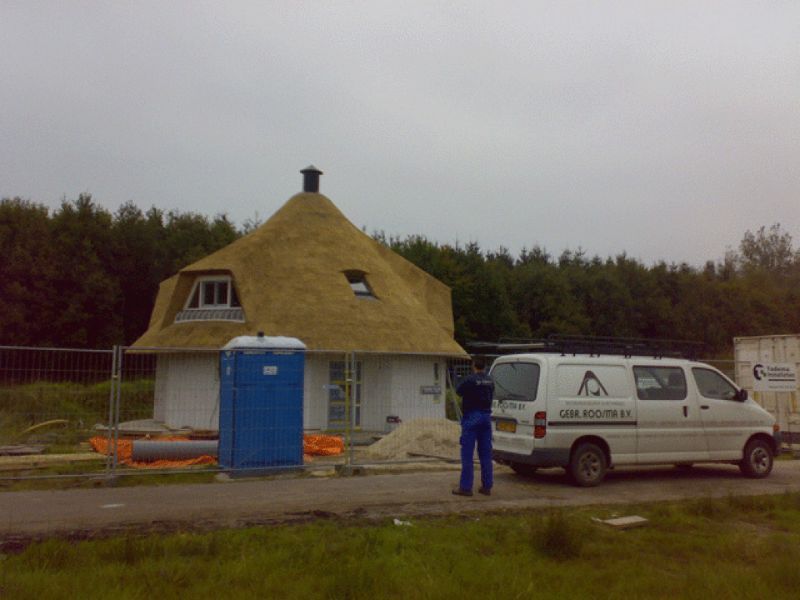 Een rond huis met een rieten dak in aanbouw. Er zijn een arbeider, een busje en een mobiel toilet aanwezig.