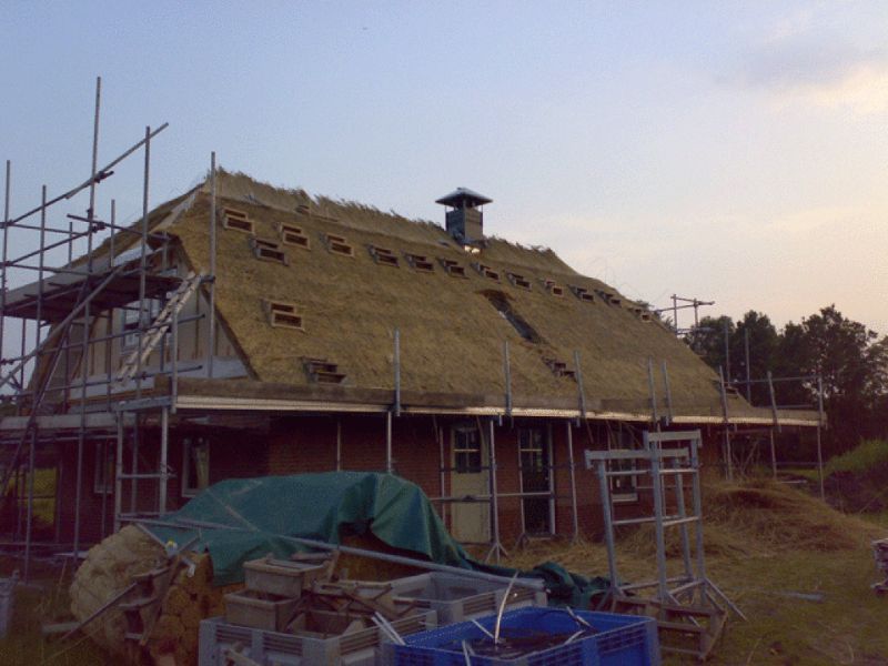 Een huis in aanbouw met een rieten dak, omgeven door steigers en bouwmaterialen buiten.