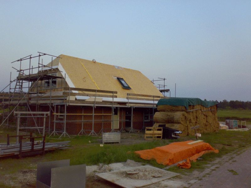 Huis in aanbouw met steigerwerk, gedeeltelijk voltooid dak en rietbalen ernaast.