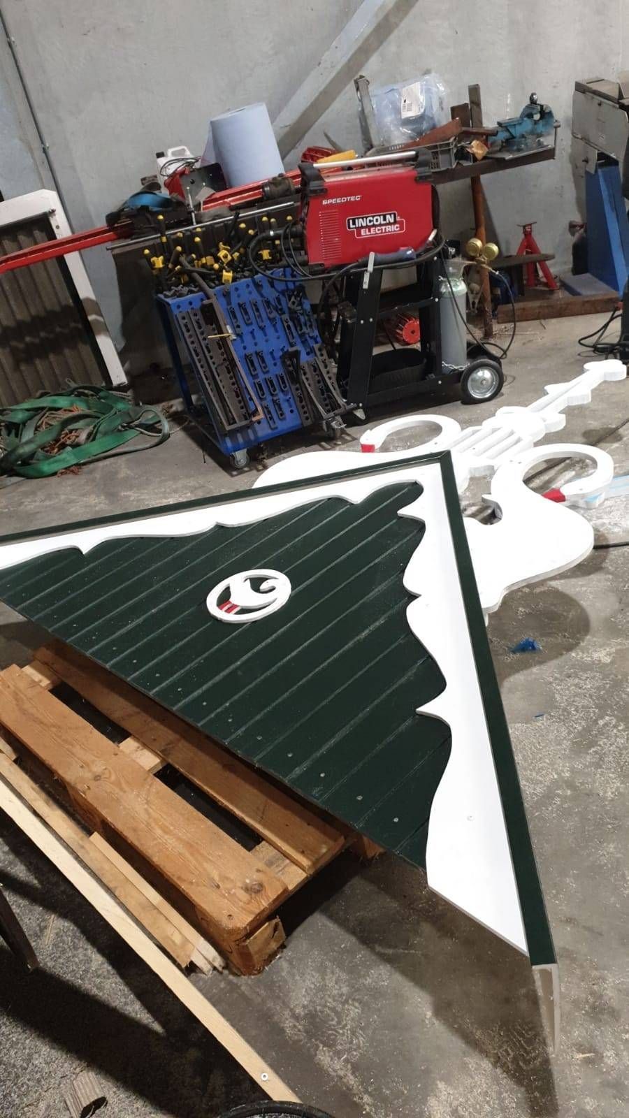 Groen-wit bord met een logo, in een rommelige werkplaats, met een pallet.