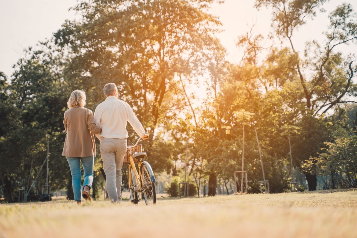 Two people walk a bicycle through a sunlit park with trees and warm evening light.
