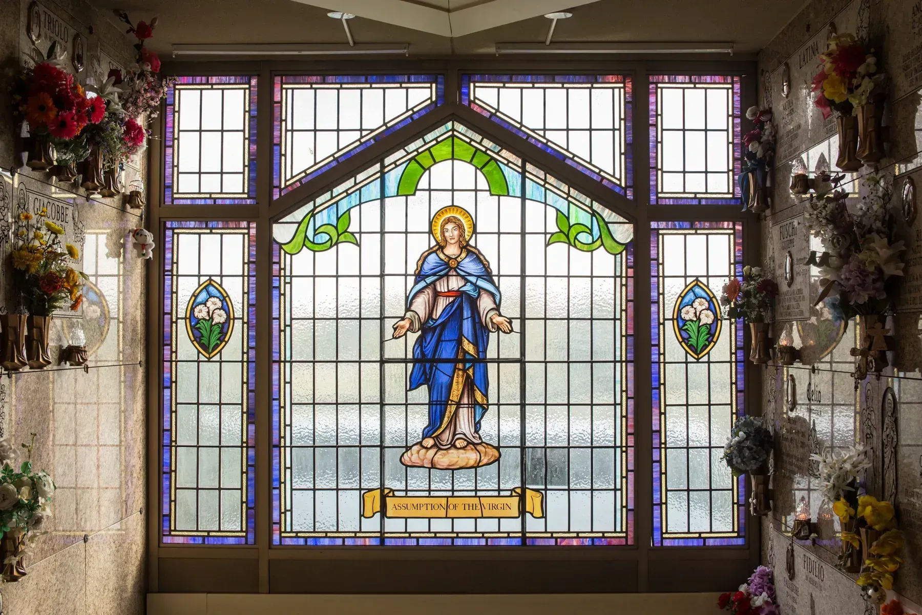 Stained glass window of a blue-robed saint holding a child, surrounded by floral decorations.